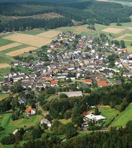 Luftaufnahme eines kleinen Dorfes umgeben von Feldern und W&auml;ldern, mit angelegten Stra&szlig;en und H&auml;usern.
