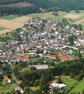 Luftaufnahme eines kleinen Dorfes umgeben von Feldern und W&auml;ldern, mit angelegten Stra&szlig;en und H&auml;usern.