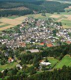 Luftaufnahme eines kleinen Dorfes umgeben von Feldern und Wäldern, mit angelegten Straßen und Häusern.