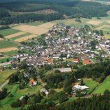 Luftaufnahme eines kleinen Dorfes umgeben von Feldern und Wäldern, mit angelegten Straßen und Häusern.