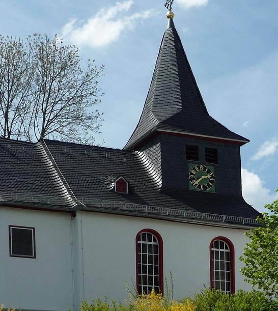 Eine evangelische Kapelle mit einem steilen Schieferdach und einem Turm in Steinbrücken. Das weiße Gebäude ist von Bäumen und Büschen umgeben und steht unter einem teilweise bewölkten Himmel.