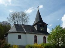Eine evangelische Kapelle mit einem steilen Schieferdach und einem Turm in Steinbrücken. Das weiße Gebäude ist von Bäumen und Büschen umgeben und steht unter einem teilweise bewölkten Himmel.