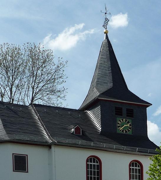 Eine evangelische Kapelle mit einem steilen Schieferdach und einem Turm in Steinbrücken. Das weiße Gebäude ist von Bäumen und Büschen umgeben und steht unter einem teilweise bewölkten Himmel.
