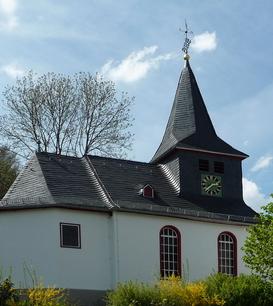 Eine evangelische Kapelle mit einem steilen Schieferdach und einem Turm in Steinbrücken. Das weiße Gebäude ist von Bäumen und Büschen umgeben und steht unter einem teilweise bewölkten Himmel.