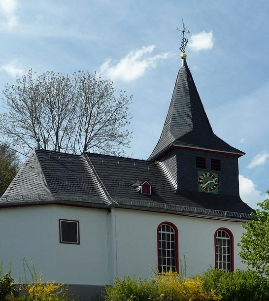 Eine evangelische Kapelle mit einem steilen Schieferdach und einem Turm in Steinbrücken. Das weiße Gebäude ist von Bäumen und Büschen umgeben und steht unter einem teilweise bewölkten Himmel.