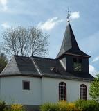 Evangelische Kapelle Steinbrücken Eine evangelische Kapelle mit einem steilen Schieferdach und einem Turm in Steinbrücken. Das weiße Gebäude ist von Bäumen und Büschen umgeben und steht unter einem teilweise bewölkten Himmel.