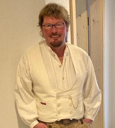 Mann mit Brille trägt eine weiße Bluse und geschnürte beige Hosen in einem hellen Raum mit Holzdetails.