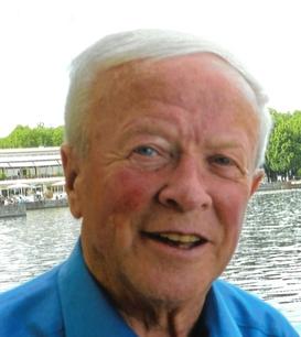 Portrait eines &auml;lteren Mannes mit kurzem, grauem Haar, l&auml;chelnd vor einem Gew&auml;sser mit gr&uuml;ner Ufervegetation.