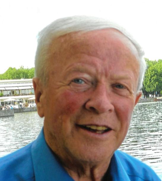 Portrait eines &auml;lteren Mannes mit kurzem, grauem Haar, l&auml;chelnd vor einem Gew&auml;sser mit gr&uuml;ner Ufervegetation.