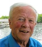 Portrait eines &auml;lteren Mannes mit kurzem, grauem Haar, l&auml;chelnd vor einem Gew&auml;sser mit gr&uuml;ner Ufervegetation.