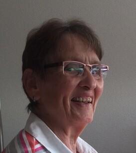 L&auml;chelnde &auml;ltere Frau mit Brille und gestreiftem Oberteil, sitzt vor einer hellen Wand.