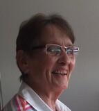 L&auml;chelnde &auml;ltere Frau mit Brille und gestreiftem Oberteil, sitzt vor einer hellen Wand.