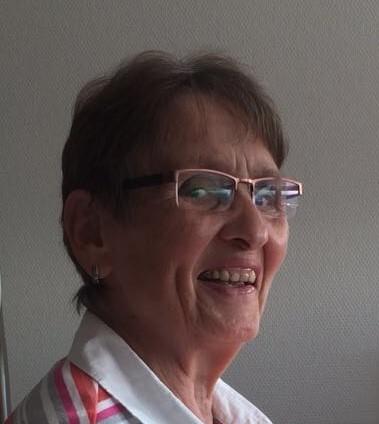 L&auml;chelnde &auml;ltere Frau mit Brille und gestreiftem Oberteil, sitzt vor einer hellen Wand.