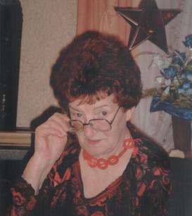 &Auml;ltere Frau mit rotem Haar tr&auml;gt eine dekorative Kette und h&auml;lt eine Brille, sitzt vor einer bl&uuml;henden Pflanze.