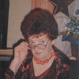 &Auml;ltere Frau mit rotem Haar tr&auml;gt eine dekorative Kette und h&auml;lt eine Brille, sitzt vor einer bl&uuml;henden Pflanze.