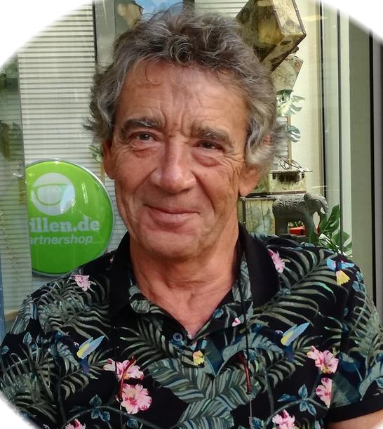 Älterer Mann mit lockigem, grauem Haar und blumigem Hemd lächelt vor einem Glasfenster mit einer grünen Werbetafel.