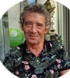 &Auml;lterer Mann mit lockigem, grauem Haar und blumigem Hemd l&auml;chelt vor einem Glasfenster mit einer gr&uuml;nen Werbetafel.