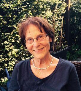 &Auml;ltere Frau mit Brille und kurzem Haar, l&auml;chelt im Freien vor einem gr&uuml;nen Hintergrund aus Pflanzen.