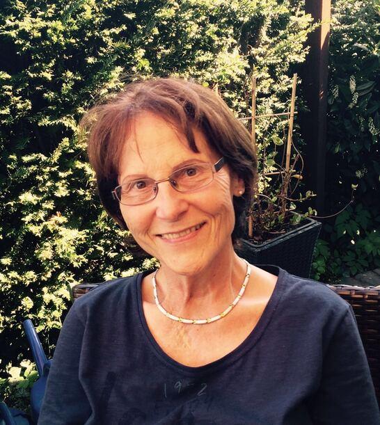 &Auml;ltere Frau mit Brille und kurzem Haar, l&auml;chelt im Freien vor einem gr&uuml;nen Hintergrund aus Pflanzen.