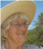 Nahaufnahme einer &auml;lteren Frau mit Sonnenhut, Brille und Graut&ouml;nen, l&auml;chelnd vor einer gr&uuml;nen Umgebung.