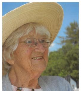 Nahaufnahme einer &auml;lteren Frau mit Sonnenhut, Brille und Graut&ouml;nen, l&auml;chelnd vor einer gr&uuml;nen Umgebung.