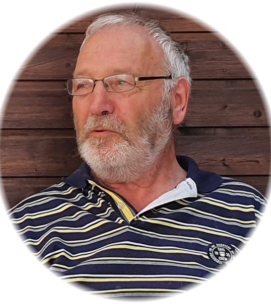 Portr&auml;t eines &auml;lteren Mannes mit Brille und grau-wei&szlig;em Bart, der in einem gestreiften Shirt vor einer Holzwand sitzt.