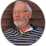 Portr&auml;t eines &auml;lteren Mannes mit Brille und grau-wei&szlig;em Bart, der in einem gestreiften Shirt vor einer Holzwand sitzt.