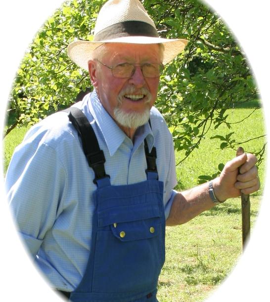 &Auml;lterer Mann mit Brille und Strohhut steht smilend im Freien, tr&auml;gt blaues Hemd und Latzhose, h&auml;lt einen Stock.