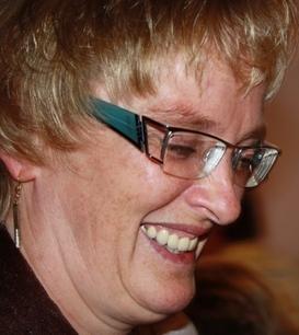 L&auml;chelnde Frau mit blonden, kurz geschnittenen Haaren und Brille, aufgenommen in einer fr&ouml;hlichen Umgebung.