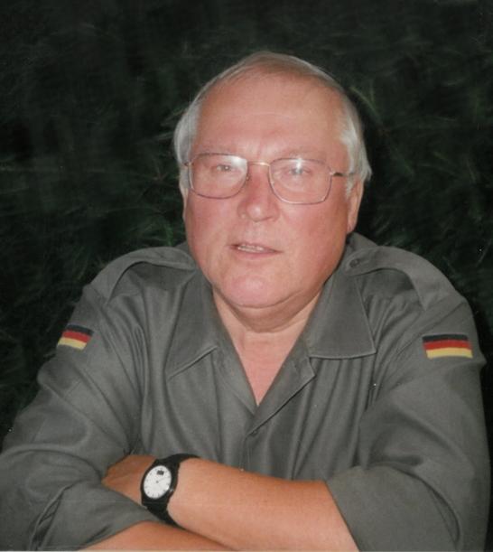 &Auml;lterer Mann mit Brille in grauer Uniform, l&auml;chelt und hat die Arme verschr&auml;nkt, Hintergrund unscharf.