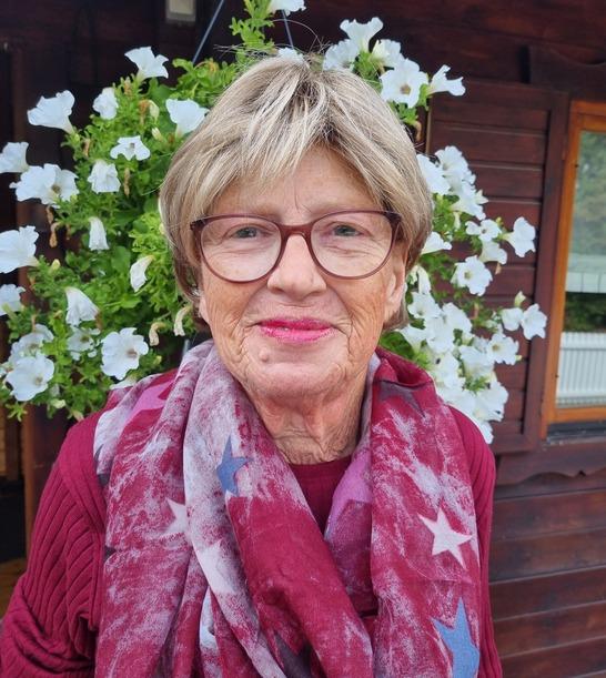 Porträt einer älteren Frau mit Brille und rotem Schal vor einer Wand aus hängenden weißen Blumen.