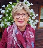 Portr&auml;t einer &auml;lteren Frau mit Brille und rotem Schal vor einer Wand aus h&auml;ngenden wei&szlig;en Blumen.