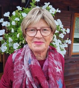 Porträt einer älteren Frau mit Brille und rotem Schal vor einer Wand aus hängenden weißen Blumen.