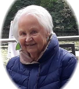 L&auml;chelnde &auml;ltere Frau mit grauen Haaren, tr&auml;gt eine blaue Jacke, sitzt an einem Tisch mit Blick auf einen See.