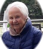 L&auml;chelnde &auml;ltere Frau mit grauen Haaren, tr&auml;gt eine blaue Jacke, sitzt an einem Tisch mit Blick auf einen See.