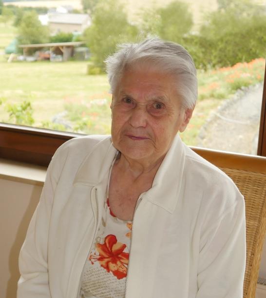 &Auml;ltere Frau mit grauen Haaren sitzt an einem Tisch, im Hintergrund eine gr&uuml;ne Landschaft und Blumen.