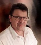 Mann mit Brille und kurzen, dunklen Haaren, tr&auml;gt ein wei&szlig;es Hemd vor einem unscharfen, dunklen Hintergrund.