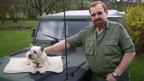 Mann im gr&uuml;nen Outfit steht neben einem Gel&auml;ndewagen, w&auml;hrend ein kleiner Hund auf der Motorhaube liegt.