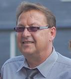 Mann mit Brille und gestreiftem Hemd l&auml;chelt vor einem neutralen Hintergrund.