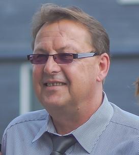 Mann mit Brille und gestreiftem Hemd l&auml;chelt vor einem neutralen Hintergrund.