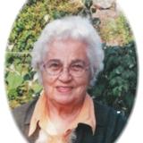 &Auml;ltere Frau mit grauem, lockigem Haar l&auml;chelt vor einer gr&uuml;nen Landschaft und h&auml;lt eine orangefarbene Katze im Arm.