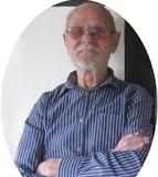 &Auml;lterer Mann mit Brille und gestreiftem Hemd l&auml;chelt, w&auml;hrend er mit verschr&auml;nkten Armen an einer Wand steht.