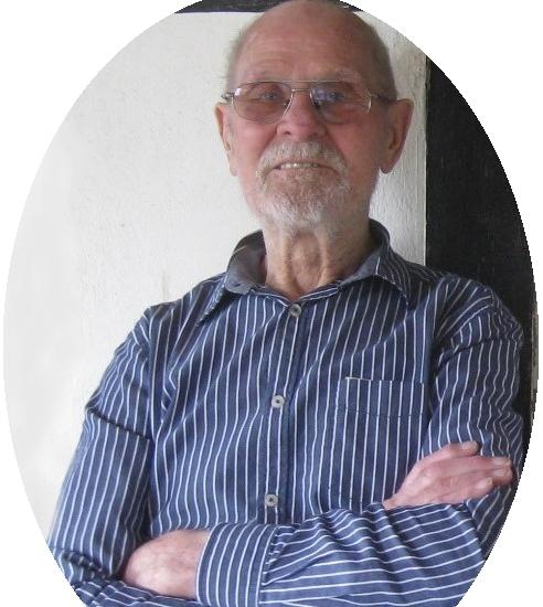 Älterer Mann mit Brille und gestreiftem Hemd lächelt, während er mit verschränkten Armen an einer Wand steht.