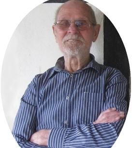 Älterer Mann mit Brille und gestreiftem Hemd lächelt, während er mit verschränkten Armen an einer Wand steht.