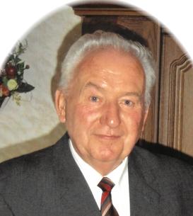 Portr&auml;t eines &auml;lteren Mannes in Anzug mit gestreifter Krawatte und einer Auszeichnung auf der linken Brust, vor Holzdekor.