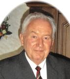 Portr&auml;t eines &auml;lteren Mannes in Anzug mit gestreifter Krawatte und einer Auszeichnung auf der linken Brust, vor Holzdekor.