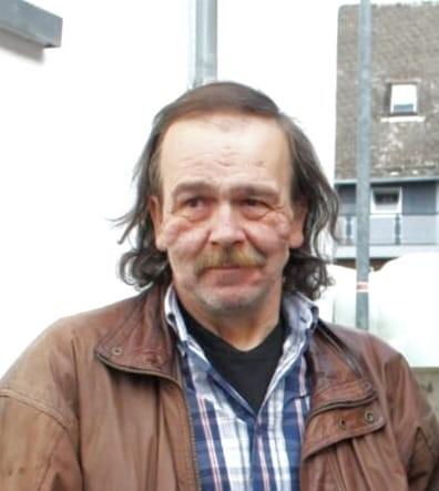 Nahaufnahme eines Mannes mit langen Haaren und einem Bart, der in einer braunen Lederjacke vor einem Geb&auml;ude steht.