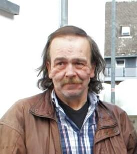 Nahaufnahme eines Mannes mit langen Haaren und einem Bart, der in einer braunen Lederjacke vor einem Geb&auml;ude steht.