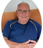 Portr&auml;t eines &auml;lteren Mannes mit Brille, der in einem blauen Shirt auf einem Sofa sitzt und l&auml;chelt.