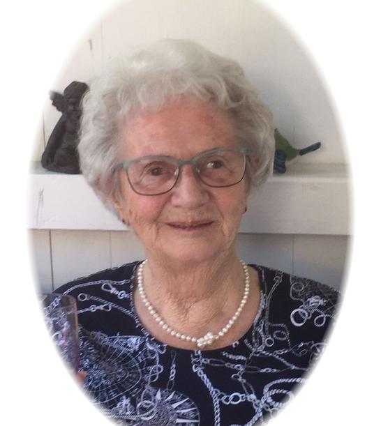 Portrait einer &auml;lteren Frau mit grauem Haar, Brille und Perlenkette vor einer hellen, schlicht dekorierten Wand.